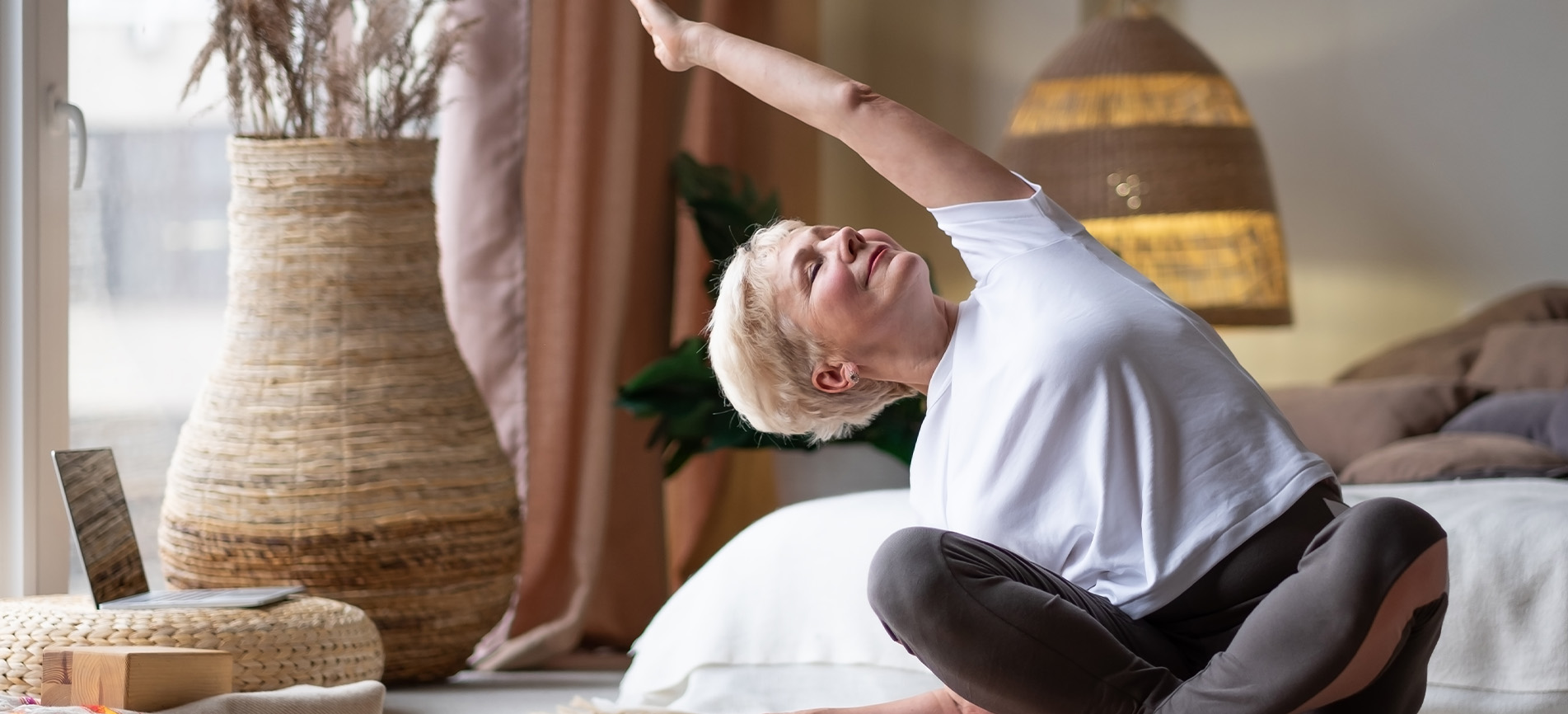 An older woman is pictured stretching upward in her sunny and bright bedroom.